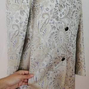 May include: A long-sleeved jacket with a paisley pattern in shades of gold, purple, and white. The jacket has a double-breasted closure with buttons.