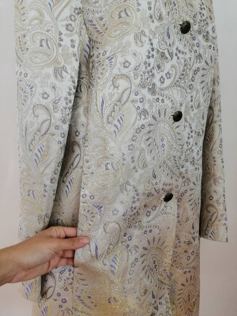 May include: A long-sleeved jacket with a paisley pattern in shades of gold, purple, and white. The jacket has a double-breasted closure with buttons.