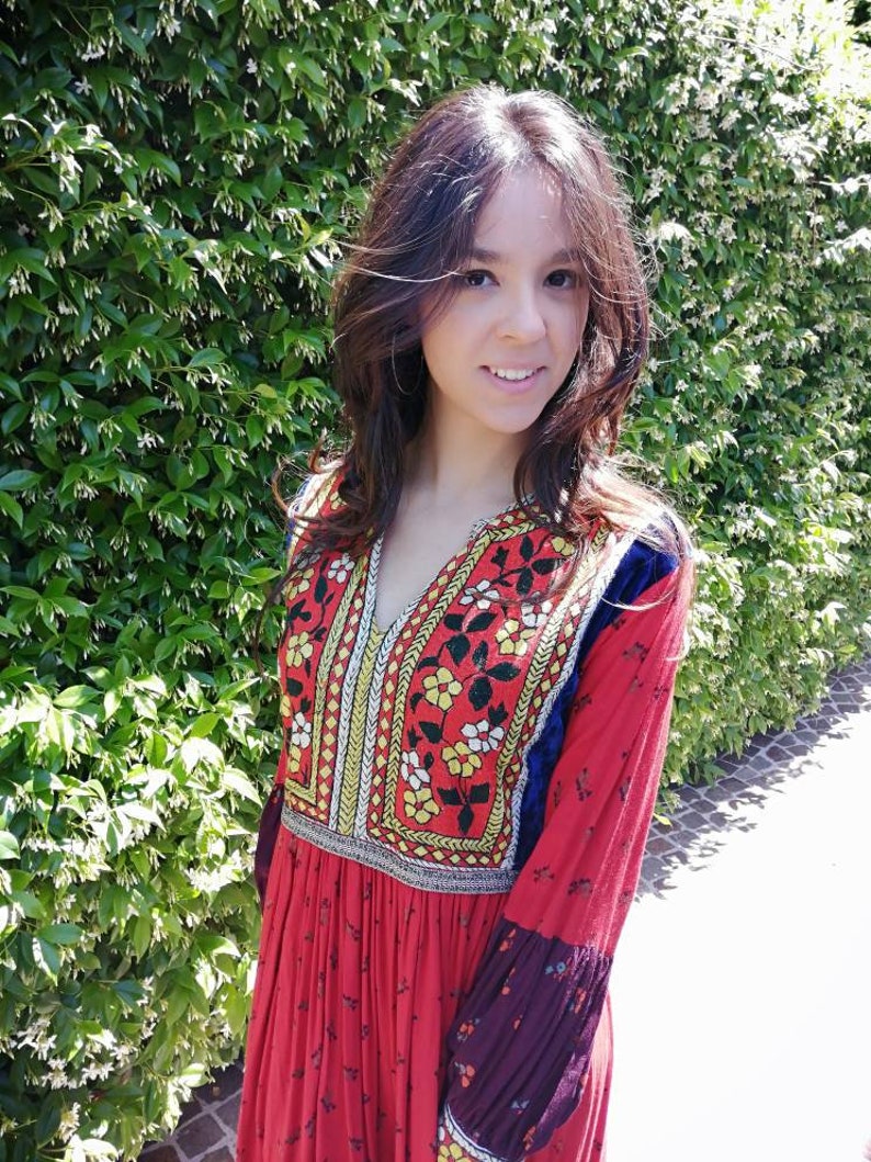Puede incluir: Una mujer con un vestido rojo de manga larga con bordados coloridos y un estampado floral. El vestido tiene un escote en V y una falda fruncida.