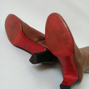 May include: A pair of brown leather heeled boots with red soles and a zipper on the back. The boots are on a white surface.