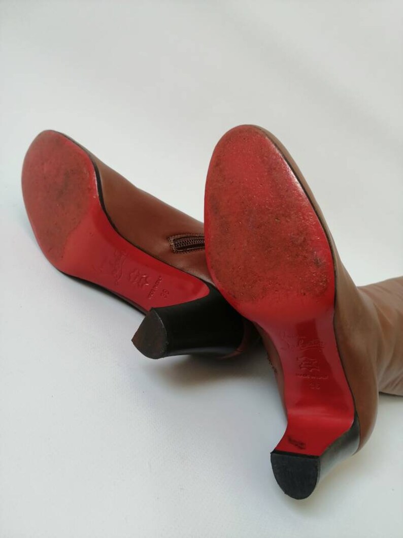 May include: A pair of brown leather heeled boots with red soles and a zipper on the back. The boots are on a white surface.