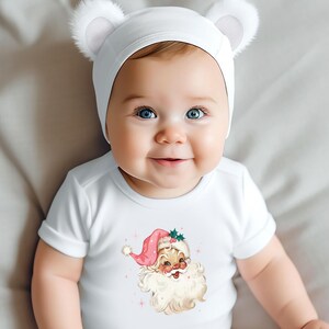 May include: A white baby bodysuit with a Santa Claus graphic, featuring a pink hat and a white beard. The baby is wearing a white hat with two small, fluffy ears.