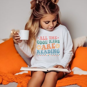 Könnte beinhalten: Ein junges Mädchen trägt ein weißes Langarmshirt mit dem Text "All the cool kids are reading" in bunten Buchstaben und einem Blumenmotiv und sitzt auf einem Sofa und liest ein Buch.