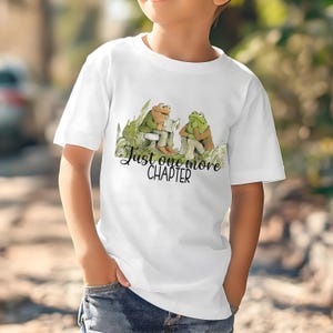 Könnte beinhalten: Weißes T-Shirt mit einer skurrilen Illustration von zwei lesenden Fröschen und dem Text "Just one more CHAPTER" in Schreibschrift. Die Frösche sind in Grün- und Brauntönen dargestellt und sitzen inmitten von grünem Laub.
