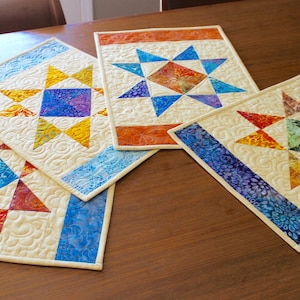 Op de afbeelding: Vier kleurrijke gequilte placemats met een stervormig ontwerp. Elke placemat heeft een ander kleurenschema, waaronder blauw, oranje, rood en groen. De placemats zijn gemaakt van een crèmekleurige stof en hebben een blauwe, oranje of paarse rand.