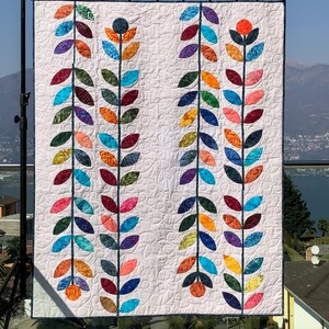 May include: A white quilt with a modern design featuring two rows of colorful leaves and flowers. The leaves are made of different fabrics and colors, including blue, green, orange, red, and purple. The flowers are simple and have a dark blue center.