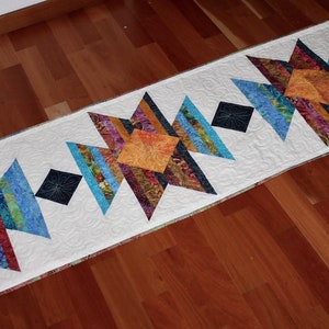 May include: A white table runner with a geometric pattern of colorful fabric squares and triangles. The pattern features a repeating design of stars with a diamond shape in the center.