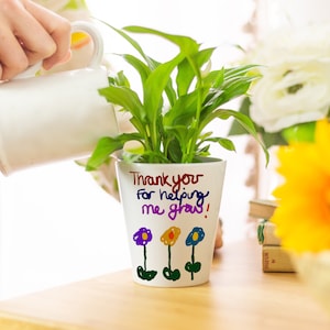 May include: White ceramic plant pot with the words "Thank you for helping me grow!" written in colourful marker. Three colourful flowers are drawn on the pot.