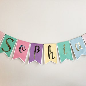 Puede incluir: Un banner colorido con el nombre "Sophia" escrito en letras doradas sobre un fondo pastel. Cada letra est&aacute; en una bandera separada con una ilustraci&oacute;n de unicornio en los extremos.