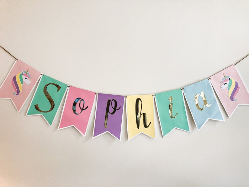 Puede incluir: Un banner colorido con el nombre "Sophia" escrito en letras doradas sobre un fondo pastel. Cada letra est&aacute; en una bandera separada con una ilustraci&oacute;n de unicornio en los extremos.
