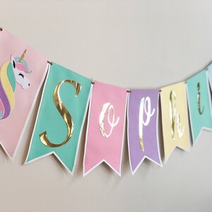 Puede incluir: Una pancarta de colores pastel con letras doradas en l&aacute;mina que deletrean "Sophia" y una ilustraci&oacute;n de unicornio. La pancarta es perfecta para una fiesta de cumplea&ntilde;os u otra celebraci&oacute;n.