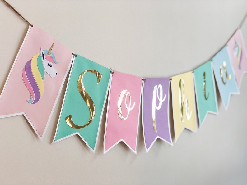 Puede incluir: Una pancarta de colores pastel con letras doradas en l&aacute;mina que deletrean "Sophia" y una ilustraci&oacute;n de unicornio. La pancarta es perfecta para una fiesta de cumplea&ntilde;os u otra celebraci&oacute;n.