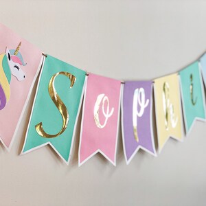 Puede incluir: Una pancarta de colores pastel con letras doradas en l&aacute;mina que deletrean "Sophia". La pancarta tiene un unicornio con una crin de arco&iacute;ris en el lado izquierdo.