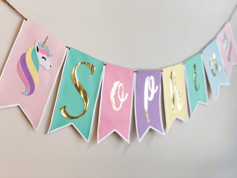Puede incluir: Una pancarta de colores pastel con letras doradas en l&aacute;mina que deletrean "Sophia". La pancarta tiene un unicornio con una crin de arco&iacute;ris en el lado izquierdo.