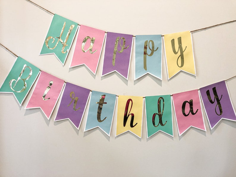 Pu&ograve; includere: Uno striscione colorato con le parole "Happy Birthday" in lettere dorate. Lo striscione &egrave; fatto di carta rosa pastello, gialla, blu e verde.