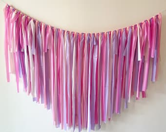May include: A pink and purple tissue paper garland with streamers hanging down. The garland is strung on a white wall.