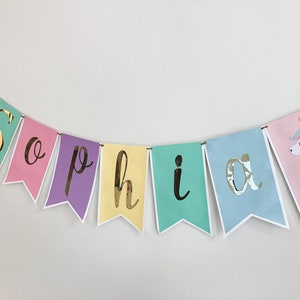 Puede incluir: Un banner colorido con el nombre "Sophia" escrito en letras doradas. Cada letra est&aacute; en una bandera de color diferente, y hay ilustraciones de unicornios en los extremos del banner.