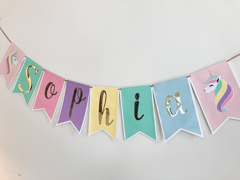 Puede incluir: Un banner colorido con el nombre "Sophia" escrito en letras doradas. Cada letra est&aacute; en una bandera de color diferente, y hay ilustraciones de unicornios en los extremos del banner.