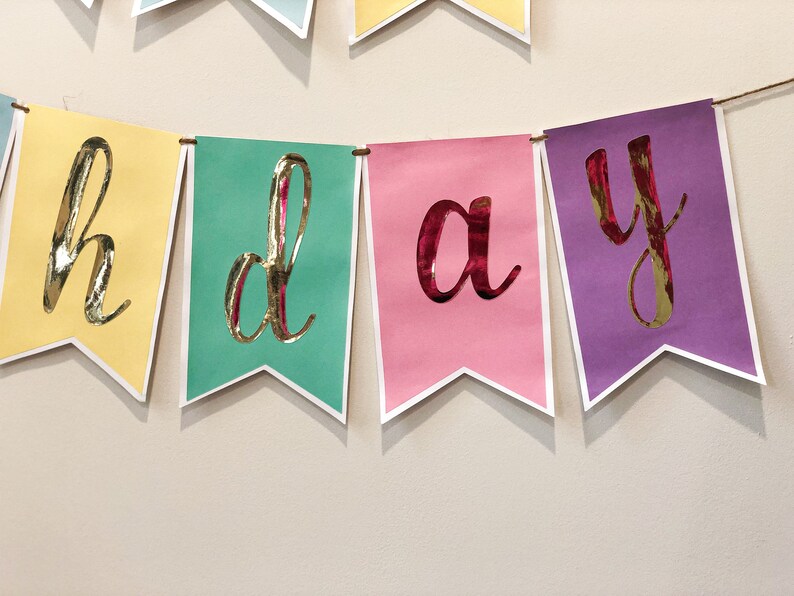 Pu&ograve; includere: Uno striscione colorato con la parola "compleanno" scritta in lettere dorate. Ogni lettera &egrave; su una bandiera separata, con le bandiere che si alternano tra giallo, verde acqua, rosa e viola.