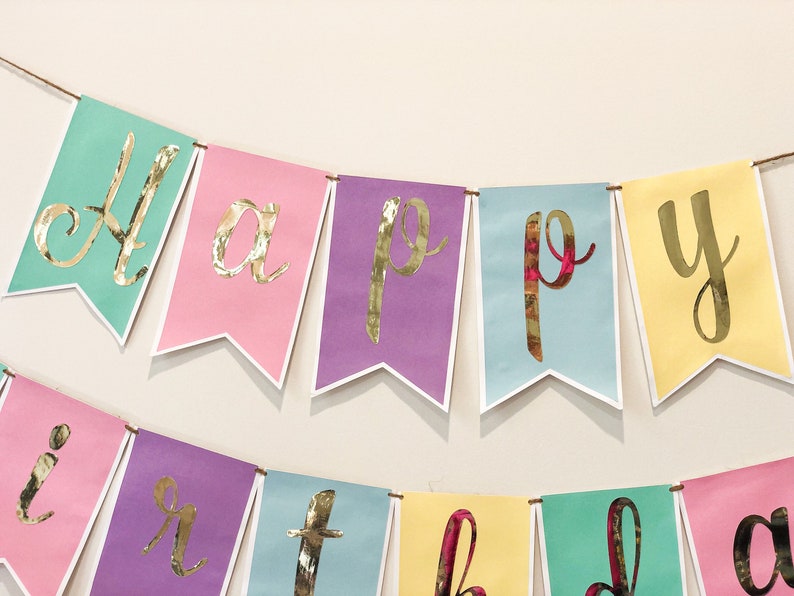 Pu&ograve; includere: Uno striscione colorato con le parole "Happy Birthday" in lettere dorate. Lo striscione &egrave; fatto di carta rosa pastello, gialla, viola, blu e verde.