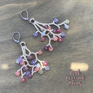 May include: A pair of beaded earrings with a floral design. The earrings feature white beads forming a branch-like structure, with clusters of pink, purple, and blue beads. The earrings have silver-coloured lever-back closures. The text "Pink Coconut Bling" is in the lower right corner.