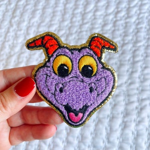 Könnte beinhalten: Lila und gold gestickter Aufnäher mit einem Cartoon-Drachen mit gelben Augen und einem rosa Mund. Der Drache hat rote Hörner und einen goldenen Glitzer-Umrand.