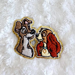 Op de afbeelding: Geborduurde patches met twee cartoonhonden, één bruin en wit, de andere bruin en rood. Beide patches hebben een gouden glitterrand en zijn geplaatst tegen een witte, donzige achtergrond.