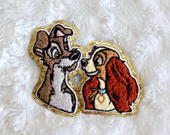 Puede incluir: Parches bordados con dos perros de dibujos animados, uno marrón y blanco, el otro marrón y rojo. Ambos parches tienen un borde dorado con purpurina y están sobre un fondo blanco y esponjoso.