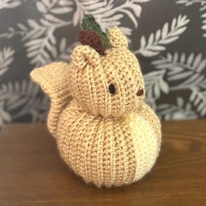 May include: A crocheted squirrel figurine in shades of tan and cream. The squirrel has a rounded body and a fluffy tail. It features small, dark eyes and a green and brown leaf detail on its head. The handmade item is displayed on a wooden surface.