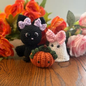 May include: Crocheted Halloween-themed toys: a black bat with a purple bow, a white ghost with a pink bow, and an orange pumpkin with green leaves. The toys are set against a backdrop of orange and pink flowers.