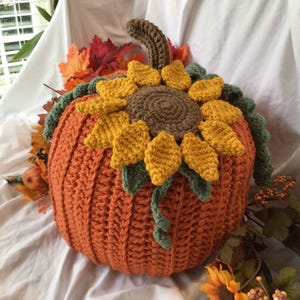 May include: A crocheted pumpkin with a sunflower on top. The pumpkin is orange and the sunflower has yellow petals and a brown center. The pumpkin is sitting on a white fabric surface.