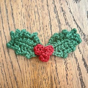 May include: A handmade crochet holly sprig with green leaves and three red berries. The leaves are detailed with textured stitches, and the berries are tightly crocheted. This festive decoration is perfect for holiday crafts and embellishments.