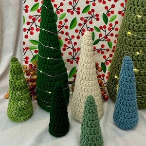 May include: Five crocheted Christmas trees in different sizes and colors. The trees are green, white, and blue. The trees are decorated with white lights.