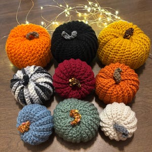 May include: A collection of nine crocheted pumpkins in various colors including orange, black, yellow, burgundy, blue, sage green, white, and a black and white striped pattern, each featuring a small textured stem on top.