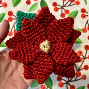 May include: A red crocheted poinsettia flower with a yellow center. The flower is held in a hand against a white background with a red berry pattern.