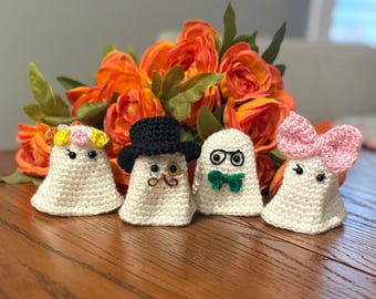 Puede incluir: Cuatro figuras de fantasmas de ganchillo hechas a mano con diferentes accesorios: una corona de flores, un sombrero de copa con monóculo, gafas y pajarita, y un lazo rosa. Los fantasmas son blancos y están sobre una superficie de madera.