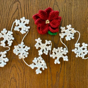 以下が含まれることがあります： 白いクロシェの雪の結晶のガーランドで、中央に赤いポインセチアの花が飾られています
