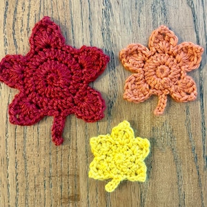 May include: Three crocheted fall leaves in shades of red, orange, and yellow. The leaves are arranged on a wooden surface. Each leaf has a detailed, textured design and a small stem.