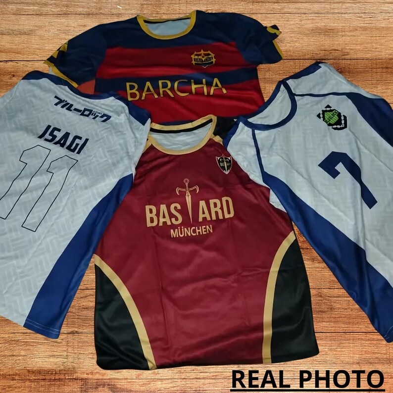May include: Three soccer jerseys with different designs. The first jersey is white with blue accents and the text "ブルーロック" and "ISAGI" on the front. The second jersey is red with gold accents and the text "BARCHA" on the front. The third jersey is white with blue accents and the text "BASTARD M&Uuml;NCHEN" on the front.