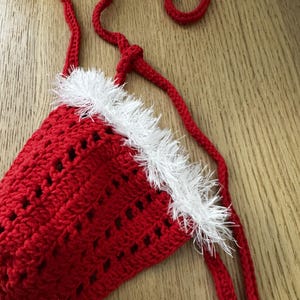 Puede incluir: Un top triangular de ganchillo rojo con un borde blanco texturizado y esponjoso en la parte superior. La prenda está confeccionada con puntos calados e incluye largos lazos de ganchillo rojo que salen de las esquinas para atar.