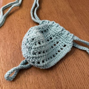 May include: A pale blue crocheted pouch with a drawstring closure. The pouch is made of a soft, textured yarn and features a rounded shape with a small, decorative element on the front. The drawstring is made of the same yarn.