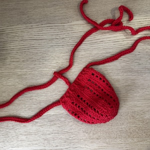 Könnte beinhalten: Ein leuchtend roter Häkelbeutel mit einem durchbrochenen Stichmuster und langen, dünnen Häkelbändern, die von den oberen Ecken ausgehen. Der Artikel ist aus Garn gefertigt und weist eine strukturierte, handgefertigte Optik mit einem mittleren Taschenbereich auf.