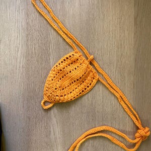 May include: A handmade, bright orange crochet bikini top. The top features a triangle shape with a textured pattern and adjustable straps. The straps are tied at the neck and back. The item is displayed on a light brown wooden surface.