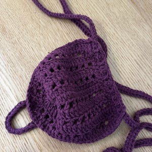 Peut inclure: Haut de bikini en crochet violet foncé fait à la main. Le haut présente une forme triangulaire avec un motif de point texturé et des bretelles réglables. Le vêtement est présenté sur une surface en bois clair.