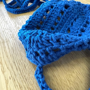 May include: A close-up of a bright blue crocheted hat with a matching strap. The hat has a textured stitch pattern and openwork design. The strap is a braided cord. The hat is handmade and appears to be made of soft yarn.