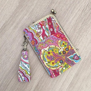 Pode incluir: Uma clutch retangular com um padrão paisley em tons de rosa, amarelo, azul e roxo. Possui um fecho de metal e uma borla a condizer. O design da bolsa é intrincado e apelativo, com uma estética vintage. A borla é presa com um fecho de metal.