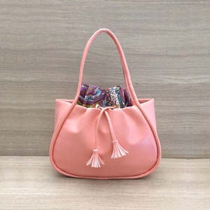 Może przedstawiać: Różowa skórzana torba typu tote z sznurkiem do ściągania i białymi frędzlami. Torba ma podszewkę w paisley.