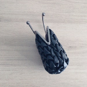 Puede incluir: Un bolso de mano negro tejido con cierre de broche plateado y asa plateada.