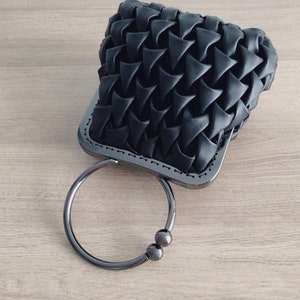 Puede incluir: Un bolso de mano negro tejido con un asa de anillo de plata y dos cuentas de plata. El bolso tiene una forma rectangular y una textura tejida.