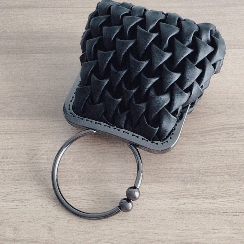 Puede incluir: Un bolso de mano negro tejido con un asa de anillo de plata y dos cuentas de plata. El bolso tiene una forma rectangular y una textura tejida.